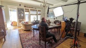 A man sits in a chair in front of a camera and laptop. He is being filmed in a living room. 