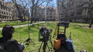 A man is being filmed outdoors in the courtyard of a university campus. A woman with dark hair sits in front of him and monitors the sound.