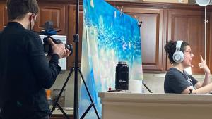 A woman stands in front of a blue cloth backdrop. She is wearing glasses and large headphones. A man stands beside her filming with a camera.