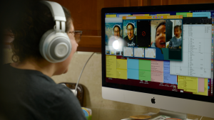 A woman sits in front of a iMac computer with large chunky headphones over her ears. On screen in front of her is a video call with two young students.