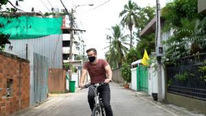 A man wearing a maroon t-shirt rides his bike through a street with a gated fenced yard on one side and a tin building on the other side.