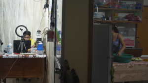 On the left side of the photo is a student in front of a draped white sheet backdrop with a ring light at his computer. The frame is split down the middle and on the right hand side is a mother preparing food in a kitchen.