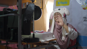 A woman sits in front of computer waving to students she teaches online. The woman wears glasses, a stuffed animal hat, and pink scarf.