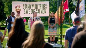 A man at a podium dressed in traditional Native American attire addresses a crowd of onlookers. Behind him a banner reads "Stand with Tribes. Save our salmon." 