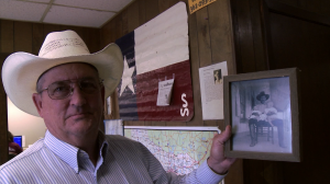 A man in a wide-brimmed cowboy had holds up a black and white photograph of a man holding two young babies. 
