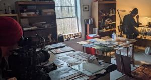 Behind the scenes photo of a busy work room, prints are strewn across multiple desks, a man sits to the right of the frame, illuminated by a yellow light, leaning over working. 