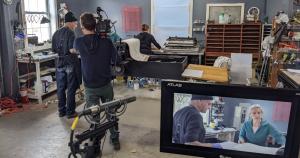In the closeup is a screen with a man and woman displayed working at a printmaking table, in the background is a cameraman filming the two and another woman working at another station in the big open studio. 