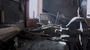 A still frame of ironworks tools. On the right side of the frame is a burning hot iron-piece that is burning hot and steaming in the light streaming in from the window. 