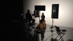 The frame is cut in half by a massive shadow. In the middle of the lighted area sits a woman in front of a white screen, speaking to a man surrounded by camera gear.