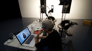 A woman sits at a computer, framed by shadow. On the computer is a picture of a woman in an editing software.