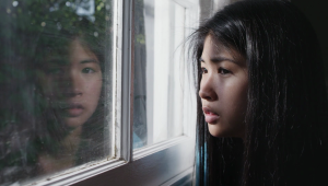 A young child with long dark hair looks out a window, her contemplative face reflects back at her.