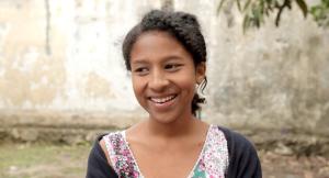 A female framed in the middle of the camera smiles off to the side of the frame, she is backlit in front of a tan wall.