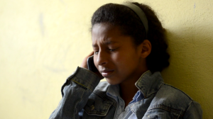 A teenage female sits, passively looking downcast. She is in shadow and the yellow wall behind her is glowing yellow.