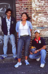 Three youth stand in front of a brick wall with grafitti. 