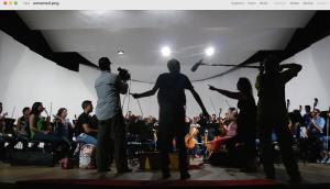 Orchestra rehearsing in theatre with camera, sound and director in front in sillhouette 