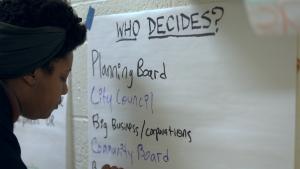 A black woman writes "Who Decides?" on a paper.