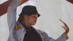 A Black nonbinary trans femme wearing a bucket hat and jean jacket vogues against a blurry bright background. They smile, gently.