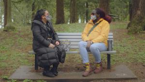 Two trans people sit on a park bench looking at one another. They wear cloth masks.