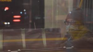 The reflection of a white transmasculine person wearing scrubs and a surgical mask looks out the window at night on an empty metro train.