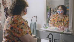 A white nonbinary person, wearing a cloth mask, rubber gloves, and brightly patterned shirt, looks in the bathroom mirror. They are surrounded by cloth masks pinned to string, drying around them.