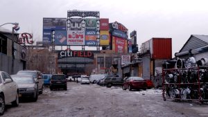 Outside the Bullpen Gate of Citi Field on a damp day with a light dusting of snow on the muddy road. Parked cars line both sides of the road. A bloated collection of corporate advertising signs hover above the stadium.
