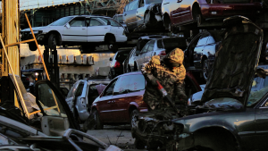 A man in an Army fatigues hoodie works on the engine of a car with a beat up front bumper in a junkyard. Behind him, cars in various states of disrepair sit on the ground or hover on platforms.