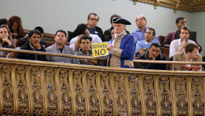 White and brown men and women sit in wooden seats behind an ornate railing. Many rest their chins wearily in their hands. An older white man in Civil War era uniform stands in front of them, glowering. He holds a sign reading "Willets NO."