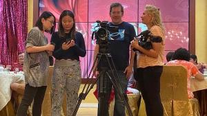 A production still for the film “Voices of Deoli” shows 4 crew members on a shoot in a restaurant, standing behind a camera on a tripod. Left of the camera, filmmaker Lulu Men looks at her phone while a female crew member looking over her shoulder. Right of the camera, cinematographer Oxana Onipko holds a camera while speaking with a man. Behind them, people sit at tables.