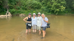 A production still for the film “Voices of Deoli” shows 4 people posing for a photograph as they stand in a brown body of water; the water reaches their calves. On the left, director Lulu Men smiles while wearing a camera strapped around her shoulder. On the right is cinematographer Oxana Onipko, also wearing a camera around her shoulder. Between them stand Deoli camp survivors Joy Ma and Michael Cheng, who are in their 60s or 70s.