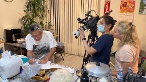 A production still from the film “Voices of Deoli” shows filmmaker Lulu Men filming a scene. She wears a face mask as she operates a camera, looking at the camera’s monitor. Cinematographer Oxana Onipko looks over her shoulder. They are filming Deoli camp survivor Bobby Wong as he signs a document on a kitchen counter crowded with cooking implements and food. A Chinese language calendar hangs on the wall behind them.