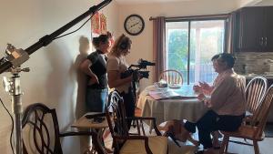 A production still from the film “Voices of Deoli” shows the filming of an interview in a home dining room. Cinematographer Oxana Onipko operates the camera while filmmaker Lulu Men stands behind her. They are filming an interview with Deoli camp survivor Andy Hsieh and his wife Jenny, who are seated at a dining room table. Andy looks up at Men, who smiles back at him. A photo album sits open on the table.