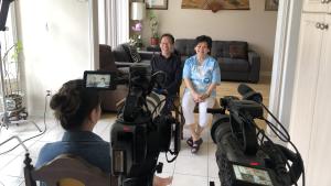 A production still from the film “Voices of Deoli” shows the filming of an interview. POV is from behind 2 cameras set up side-by-side, filming Deoli camp survivor Moses Cheng and his wife Sherry, who sit in chairs in a living room. They smile at the cameras. A monitor on the left camera shows a medium shot of the couple. Filmmaker Lulu Men sits left of the 2 cameras; only the back of her head is visible. 