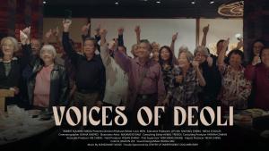 A horizontal format poster for the film “Voices of Deoli” features a group of approximately 20 seniors who are Deoli camp survivors. They are in a restaurant. They smile and each raise one arm with a thumbs up. They look to the left, offscreen. The bottom of the poster features the title of the film and various credits.