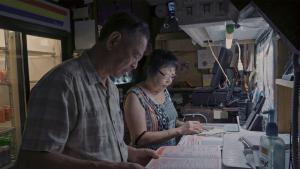 A production still from the film “Voices of Deoli” shows Deoli camp survivor Michael Cheng and his wife Patsy looking at papers in an office or workshop space. Patsy wears glasses and holds a pen, looking down at a paper, while Michael looks at a menu. There are 2 landline phones and a computer in the background.