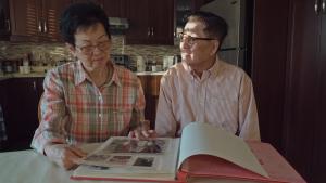 A production still from the film “Voices of Deoli” shows Deoli camp survivor Andy Hsieh and his wife Jenny sitting side-by-side at a kitchen table. They both wear glasses. Jenny looks down at an open photo album on the table while Andy turns his face to look at her. His face is covered in shadows created by sunlight pouring through a nearby kitchen blind.