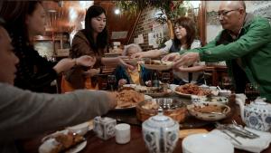 A production still from the film “Voices of Deoli” shows a family sharing a meal at a table. They hand plates of food to each other across the table. Pictured are Stephanie Ma, a young woman who stands next to Grand Aunty, who is seated. On the right are Joy Ma and William Ma, both Deoli camp survivors. Two additional people are on the left. Several large bowls of food and teapots sit on the table.