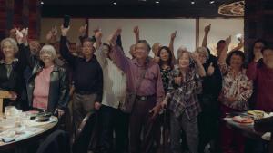 A production still from the film “Voices of Deoli” features a group of approximately 20 seniors who are Deoli camp survivors. They are in a restaurant. They smile and each raise one arm with a thumbs up. They look to the left, offscreen.
