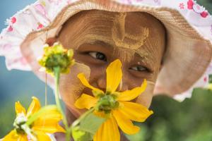 A bald child with cancer with yellow face paint is wearing a sunhat and smelling a yellow flower, while looking off to the right.  