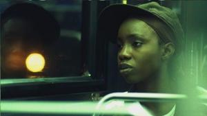 A close up image of a Black youth sitting in a bus with their face reflected on the bus window as they peer out into the distance. A harsh white street light obscures their reflection against the dark night background.