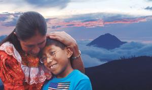In Guatemala, a woman cradles the head of a young person with sunset-red clouds and a tall mountain in the distance behind them. They are both smiling. The woman wears a red traditional Guatemalan dress and the boy wears a blue t-shirt with a U.S. flag printed on it.