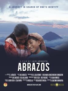 In Guatemala, a woman cradles the head of a young person with sunset-red clouds and a tall mountain in the distance behind them. They are both smiling. The woman wears a red traditional Guatemalan dress and the boy wears a blue t-shirt with a U.S. flag printed on it. Text, "Abrazos. A journey in search of one's identity. A film by Luis Argueta."