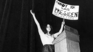 In this black and white still from the film, a young woman triumphantly holds up a sign that reads “Sandy For Class President.” The word ‘President’ is crossed out and underneath is written “Secretary”. She’s onstage at a lectern.