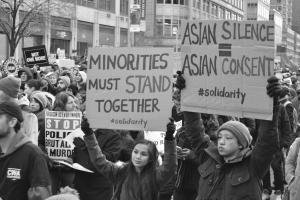Black and white photo of Asian-American protesters holding signs. One reads Minorities must stand together.  Another sign reads Asian silence equals asian consent.