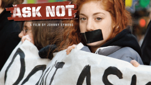 A young girl with red hair stares boldly into the camera in a protest. She has a large black strip of duct tape covering her mouth and she is holding up a protest banner. The other people holding up the banner are blurred in the background. Film title, "Ask Not."