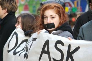 A young girl with red hair stares boldly into the camera in a protest. She has a large black strip of duct tape covering her mouth and she is holding up a protest banner. The other people holding up the banner are blurred in the background.