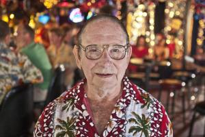 A still from the New Day Film Before You Know It. An balding, elderly man with glasses and a brightly patterned button down shirt looks into the camera with an amused grin. People and bright colored lights fill the blurred background.