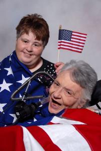 A still from the New Day Film Body & Soul: Diana and Kathy. Two women lean into each other, with the red haired woman’s cheek pressing against the side of the gray haired woman’s forehead. The red haired woman is wearing a blue shirt with white stars and the gray haired woman wears a red shirt. They both look towards the camera and smile.