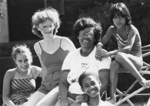 In this black and white photo from the 1980s, five teens and children of various races sit on playground equipment smiling on a bright summer’s day.