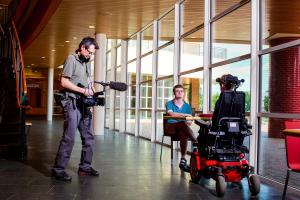 Filmmaker Robert Rooy is standing in a building and is pointing a video camera at two people sitting at a table. One of the people is in a wheel chair. 