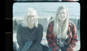 A woman with medium blond hair with her daughter with long blond hair sitting in front of ocean waves